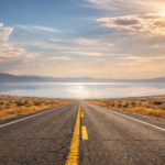 Ruta recta en un paisaje árido con un espejismo que simula un lago en el horizonte bajo un cielo con nubes y luz dorada.